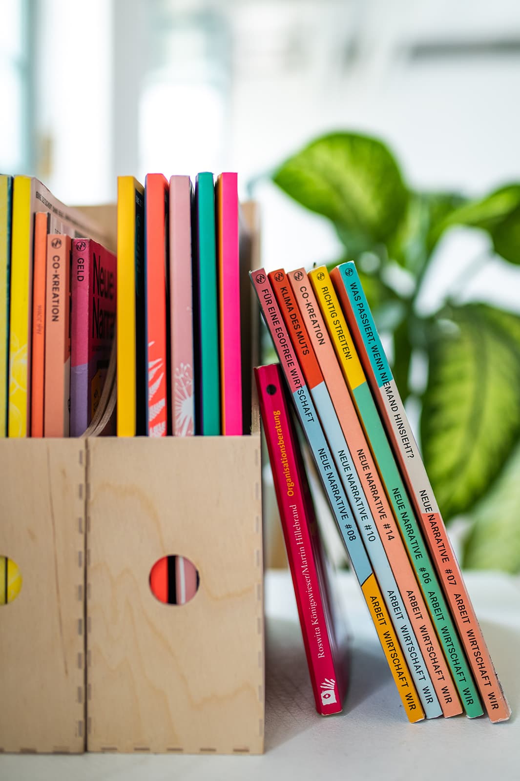 Bunte Bücherstapel in Holzbox auf Schreibtisch mit Pflanzen im Hintergrund.