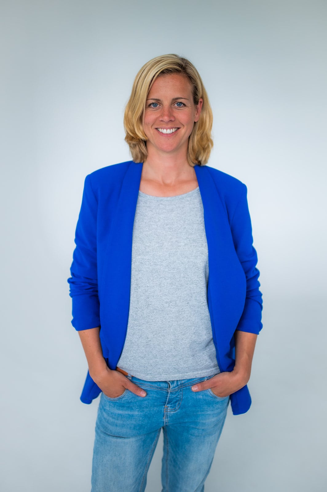 Frau in blauer Jacke und Jeans, lächelnd, vor neutralem Hintergrund.