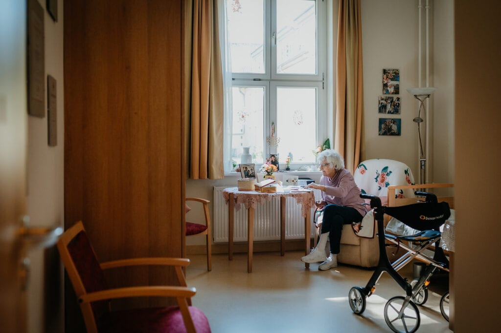 Ältere Frau beim Lesen im hellen Wohnzimmer mit großem Fenster und Pflegebett.