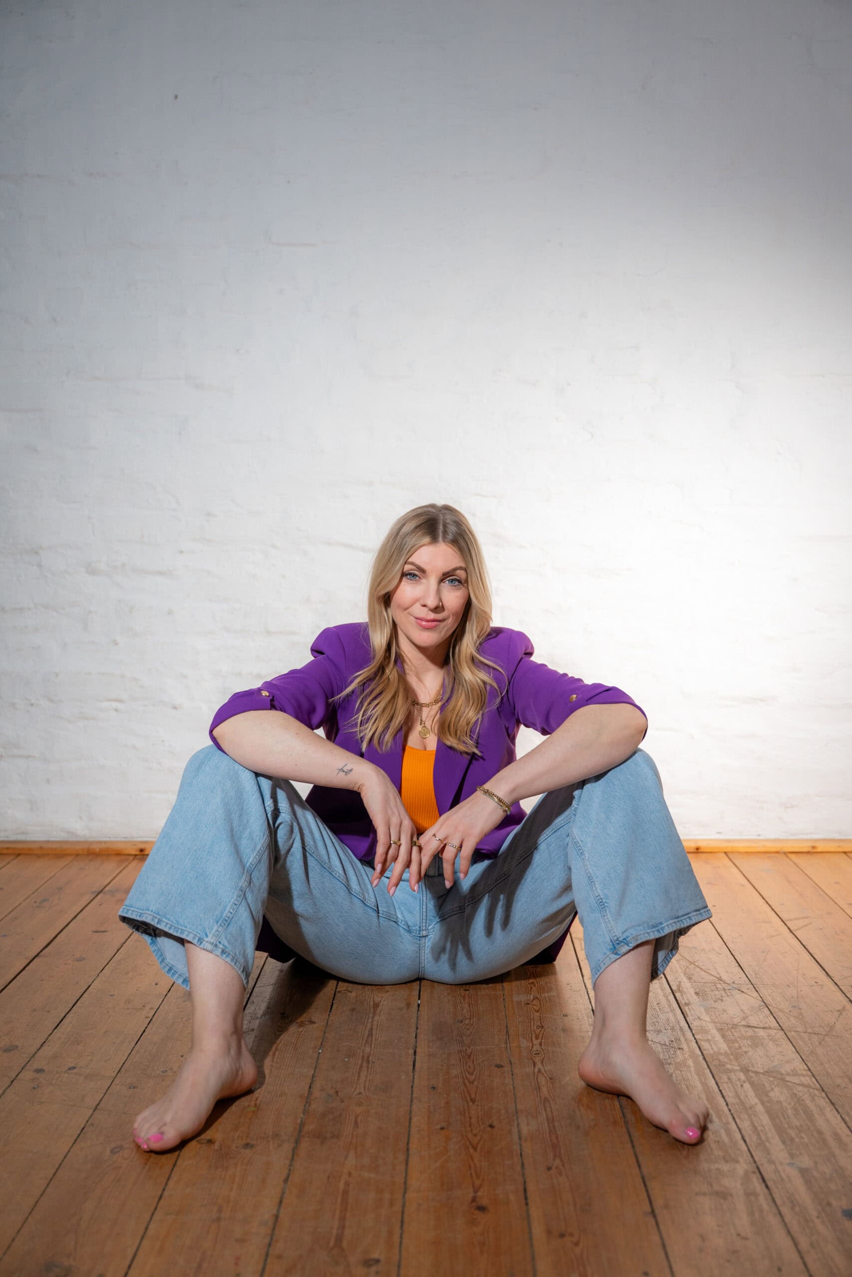 Selbstbewusste Frau in buntem Outfit sitzt entspannt auf Holzboden vor weißer Wand.