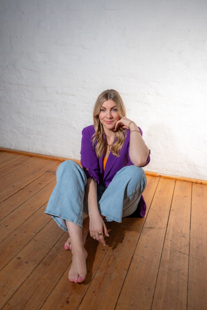 Frau sitzt auf Holzboden, trägt lila Blazer und Jeans, lächelt freundlich.