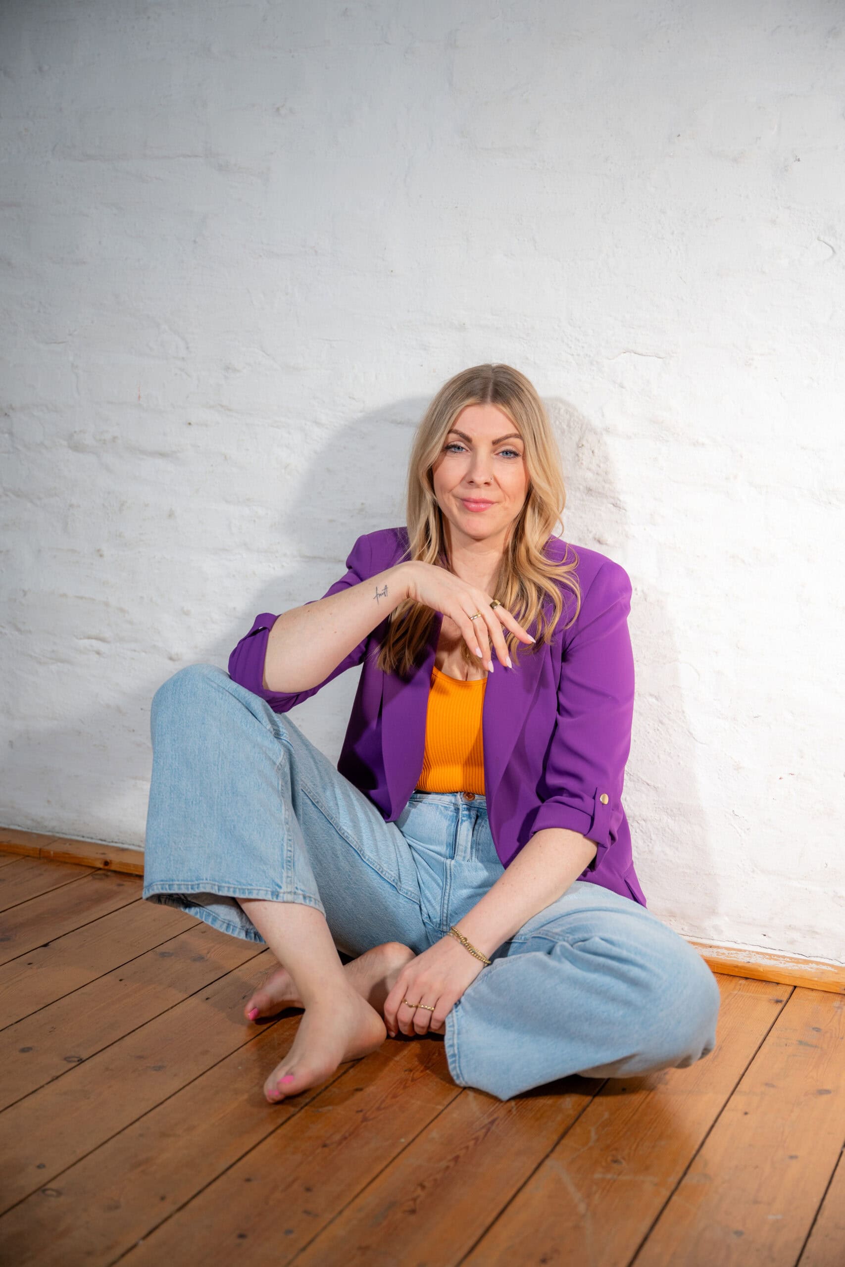 Frau in buntem Outfit sitzt auf Holzboden vor weißer Wand.