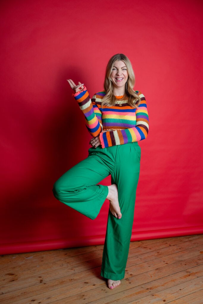 Bunte Frau in farbenfrohem Outfit macht Yoga-Pose vor rotem Hintergrund.