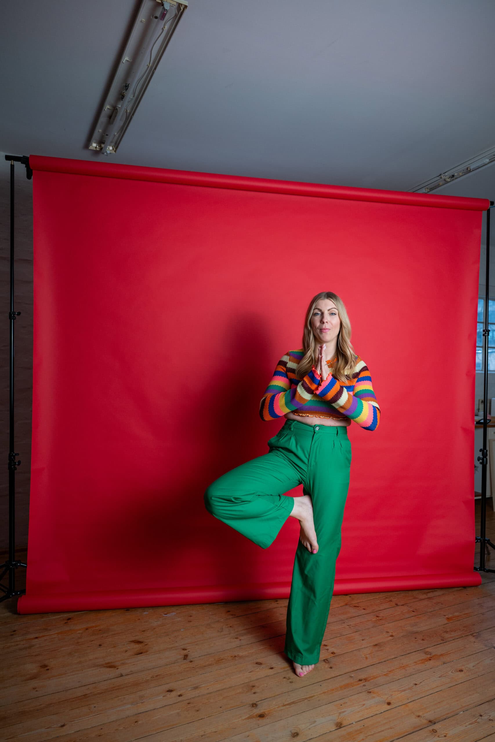 Frau in buntem Pullover und grünen Hosen macht Yoga-Übung vor rotem Fotohintergrund.