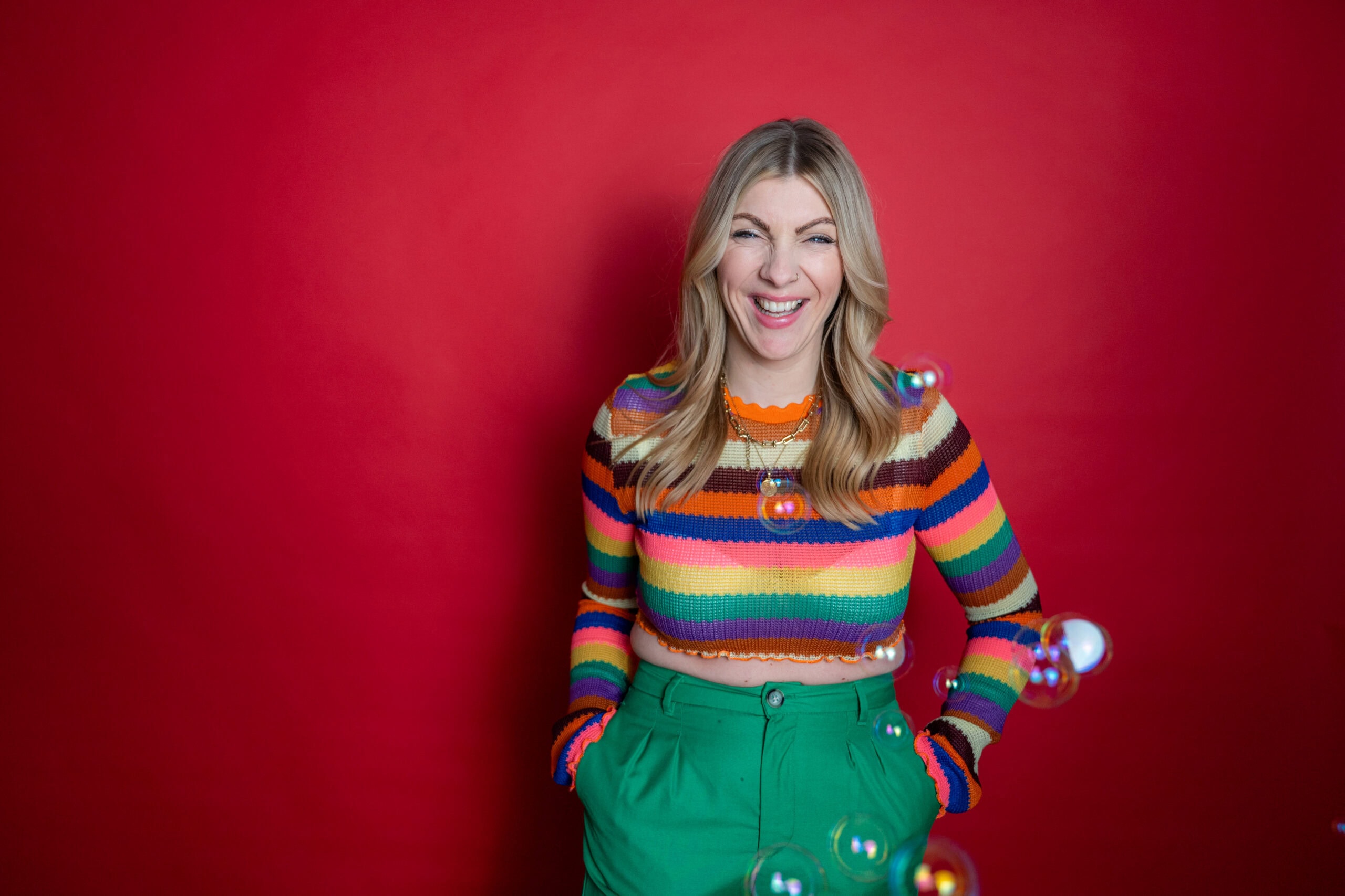 Fröhliches Mädchen in buntem Pullover vor rotem Hintergrund, lachend und spielend mit Seifenblasen.