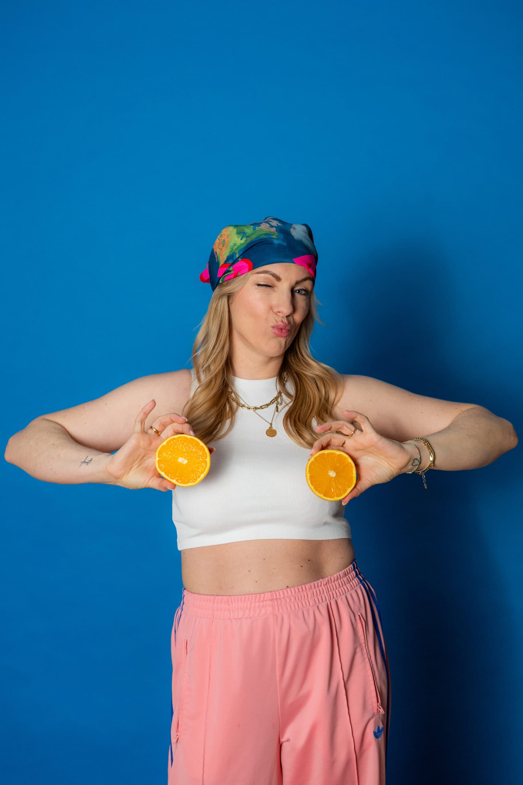 Fröhliche Frau mit buntem Kopftuch hält Orangen vor der Brust, lachend und entspannt vor blauem Hint.