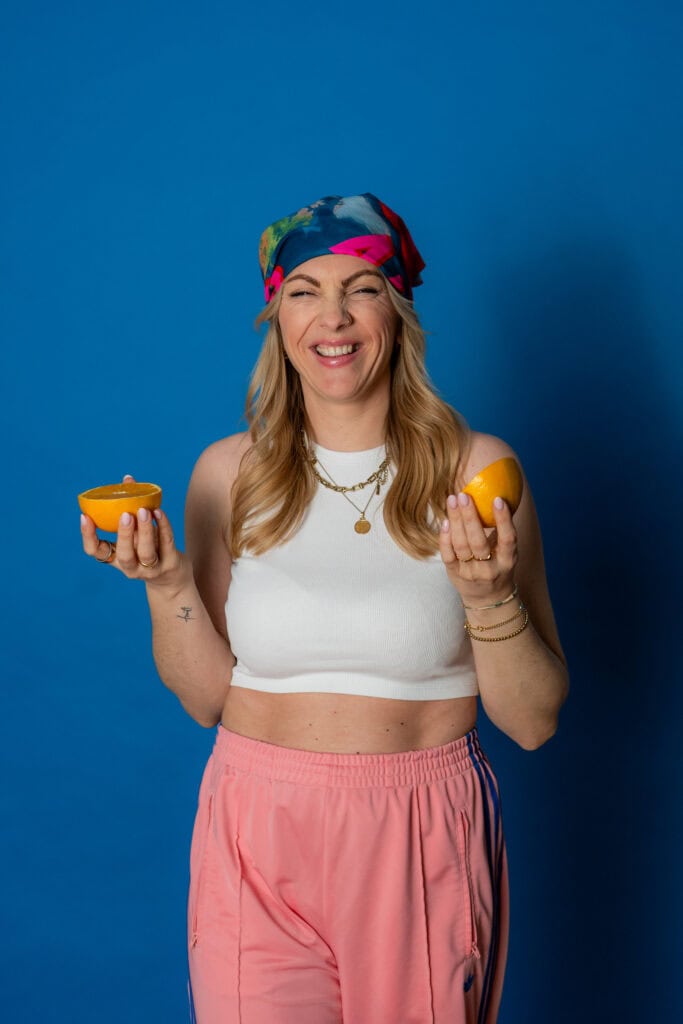 Fröhliche Frau hält Zitrusfrüchte, trägt bunte Kopfbedeckung und weiße Top, steht vor blauem Hinterg.