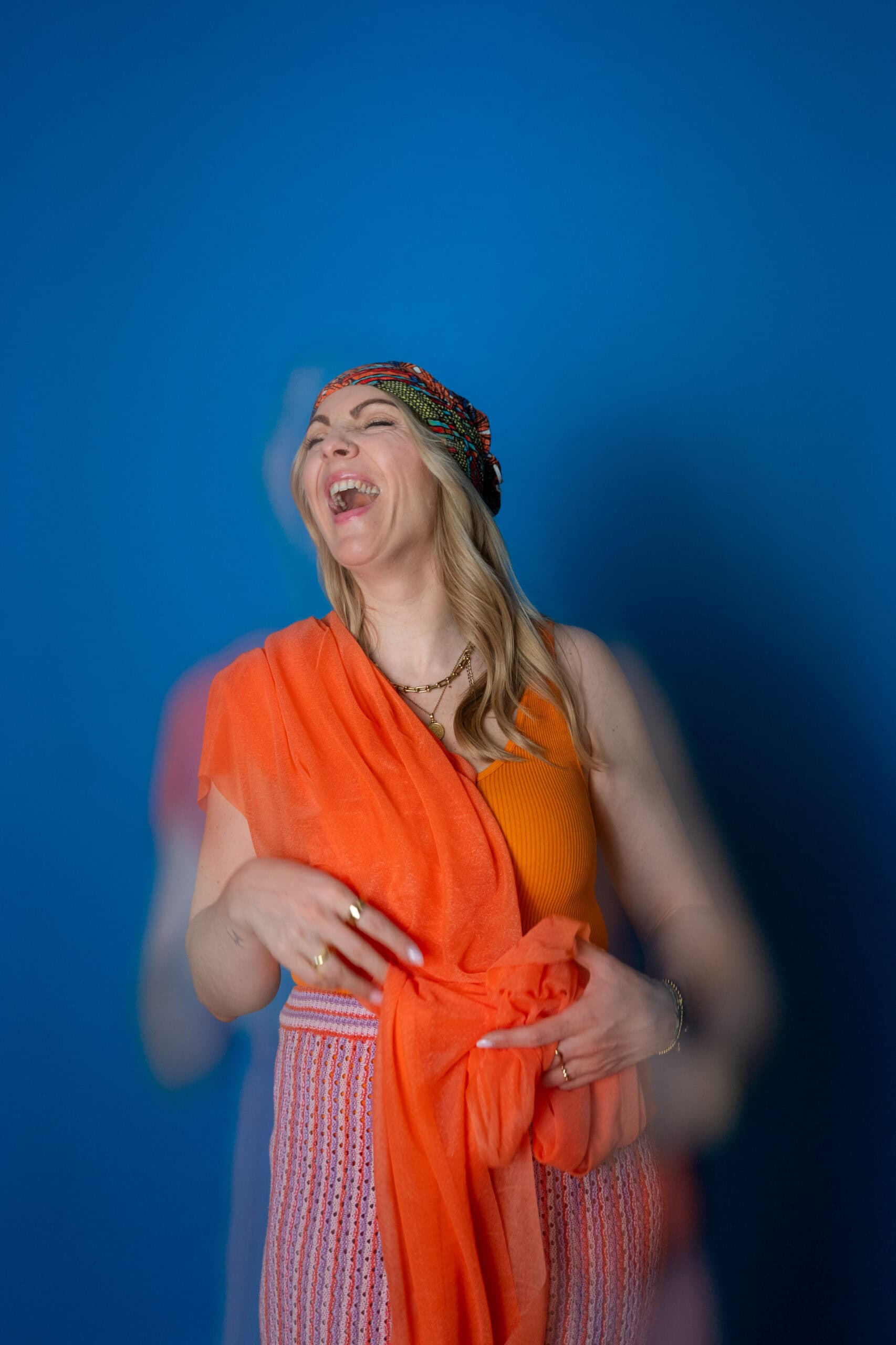 Frau lacht herzlich vor einem blauen Hintergrund, trägt ein buntes Kopftuch und ein orangefarbenes K.