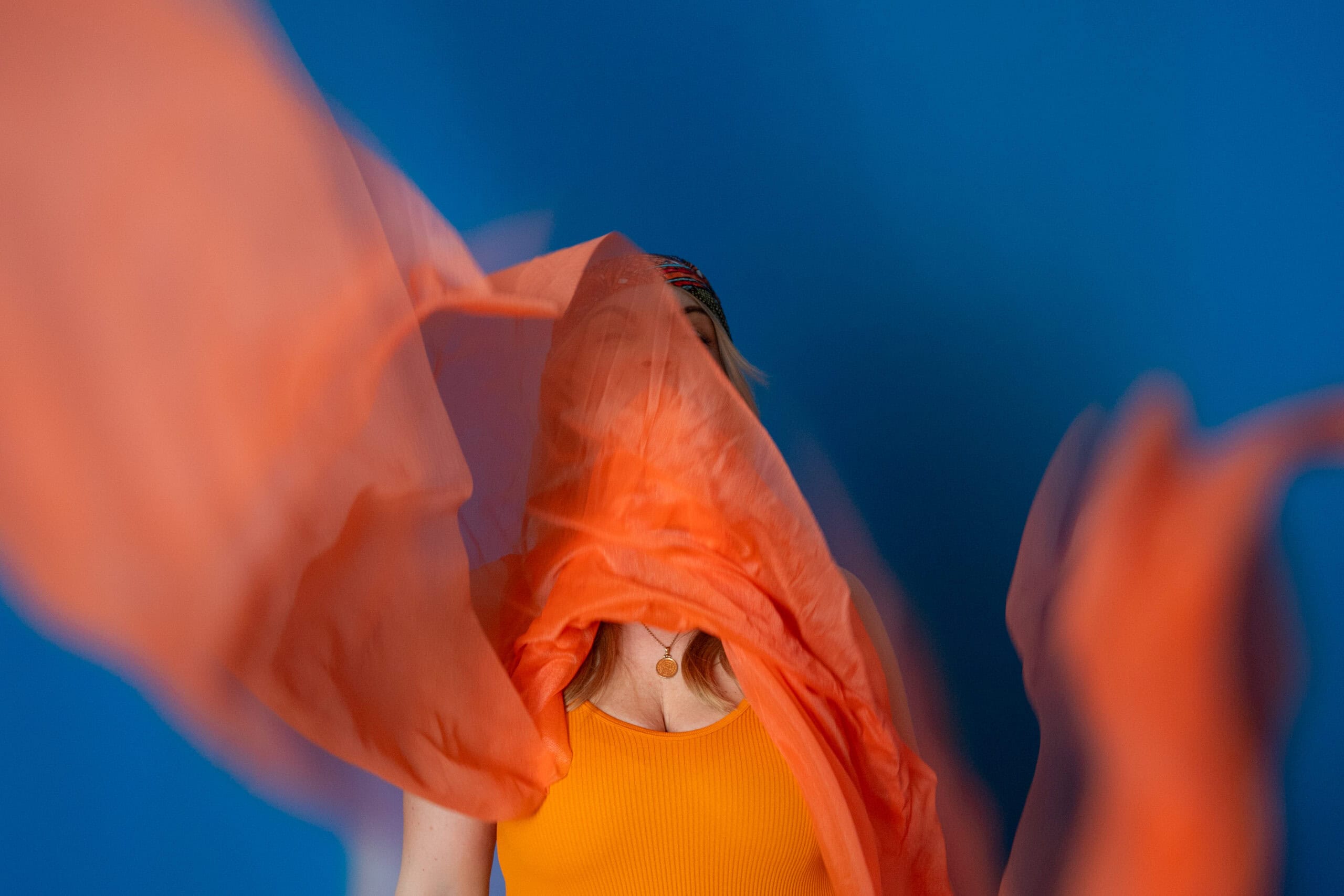 Künstlerische Fotografie mit fließendem orangefarbenem Stoff vor einer blauen Wand.