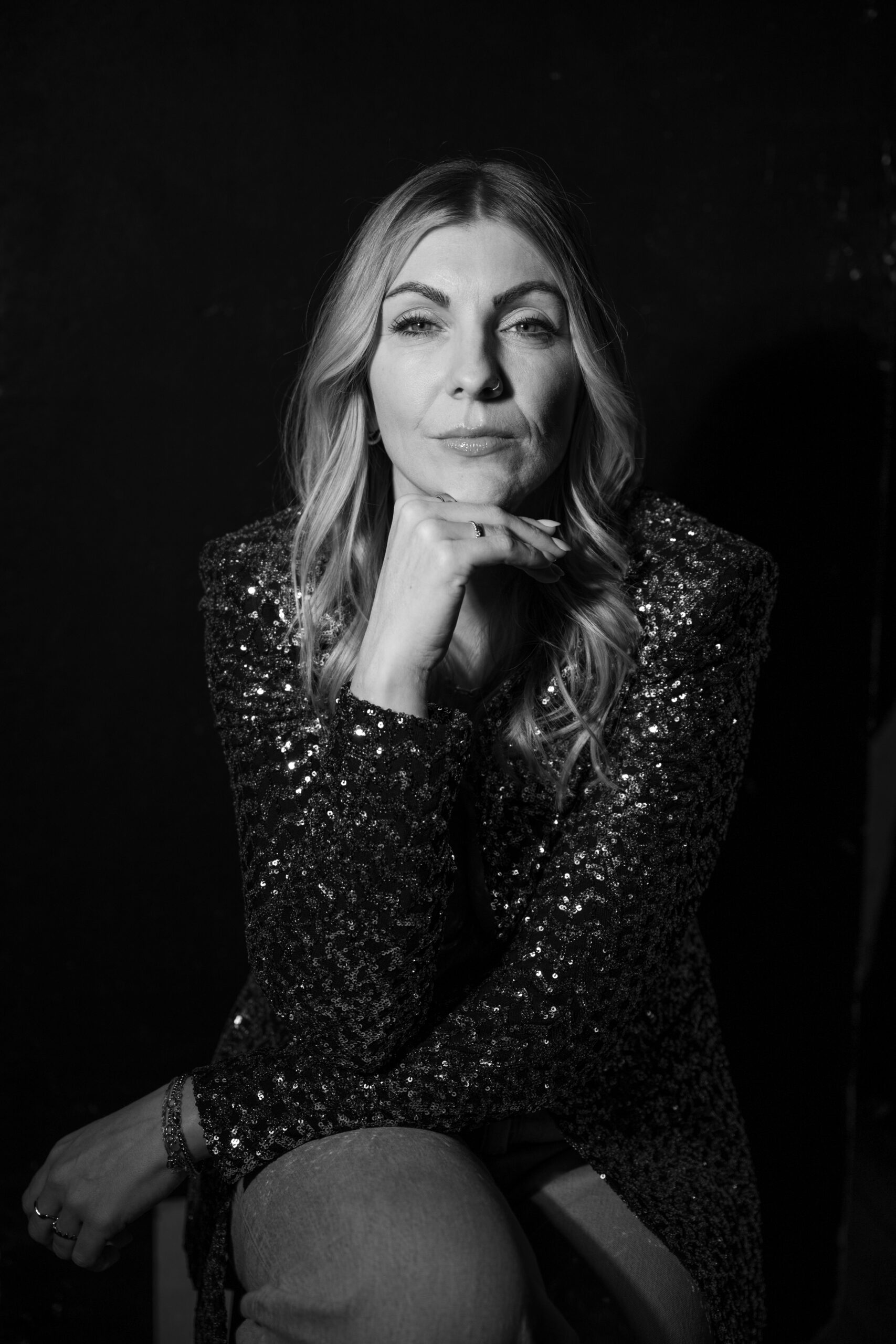 Frau in Glitzerjacke sitzt nachdenklich, schwarz-weiß, elegante Pose.