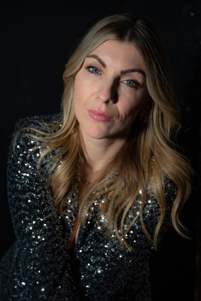 Frau mit blonden Haaren und Glitzer-Outfit, blickt selbstbewusst in die Kamera.