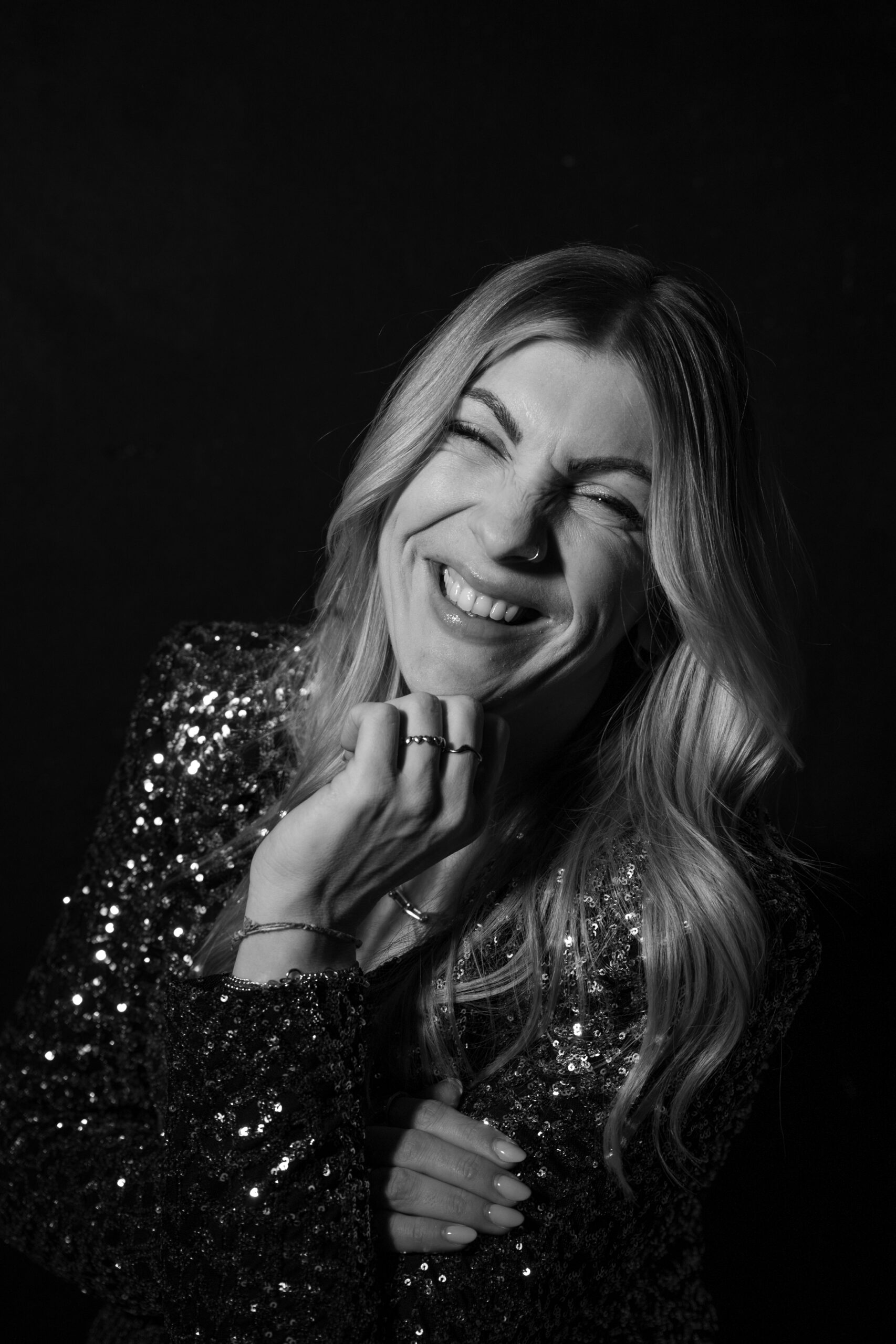 Frau lacht fröhlich, trägt eine funkelnde Jacke, schwarze Hintergrund, positive Stimmung.