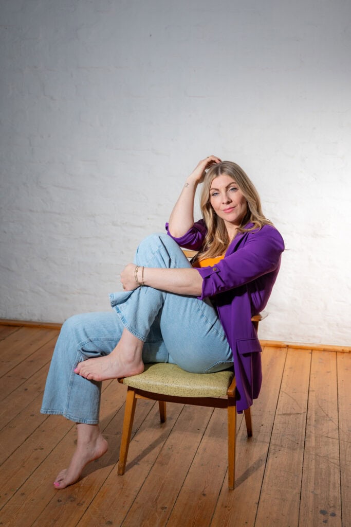 Junge Frau in buntem Outfit sitzt auf Stuhl vor weißer Wand.