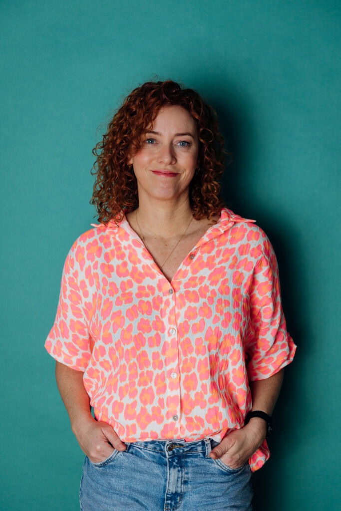 Frau mit lockigem Haar trägt farbiges Hemd, Portrait vor grünem Hintergrund.