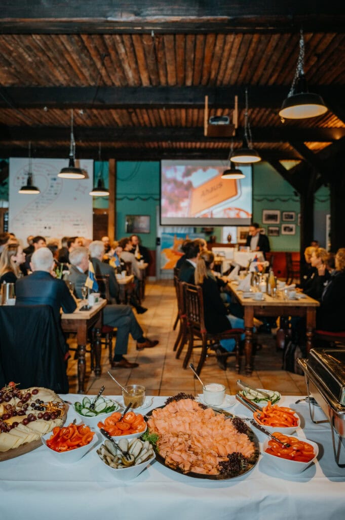 Veranstaltungsraum mit Publikum und Buffet, Präsentation im Hintergrund.