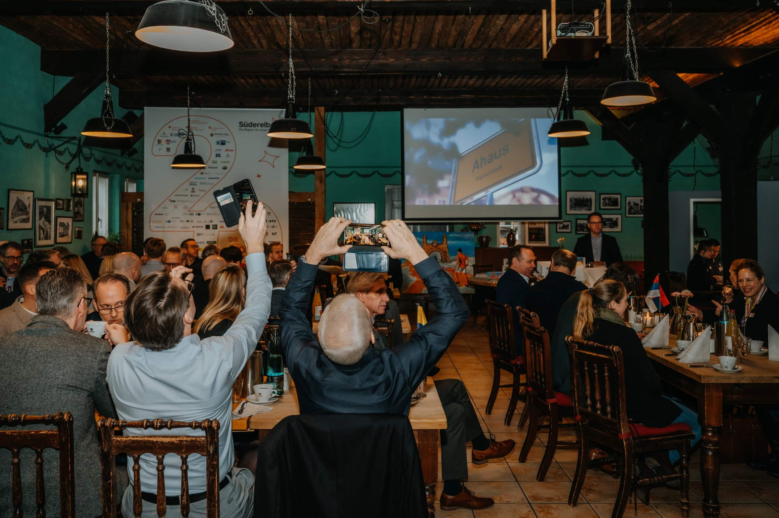 Menschen bei einer Veranstaltung im rustikalen Veranstaltungsraum, viele nehmen Fotos auf.
