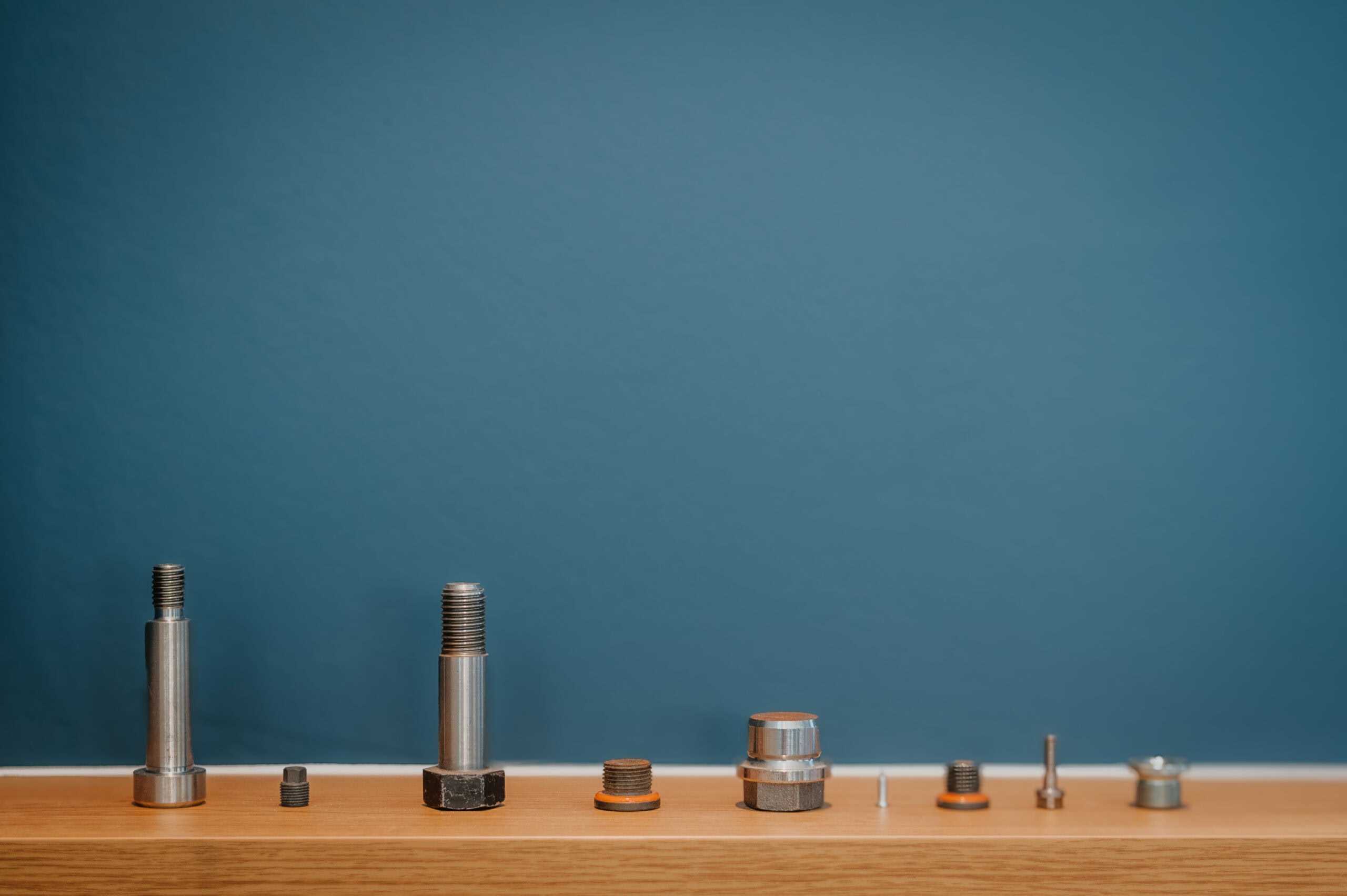 Verschiedene Schrauben und Muttern in einer Reihe auf Holz, mit blauer Wand im Hintergrund.