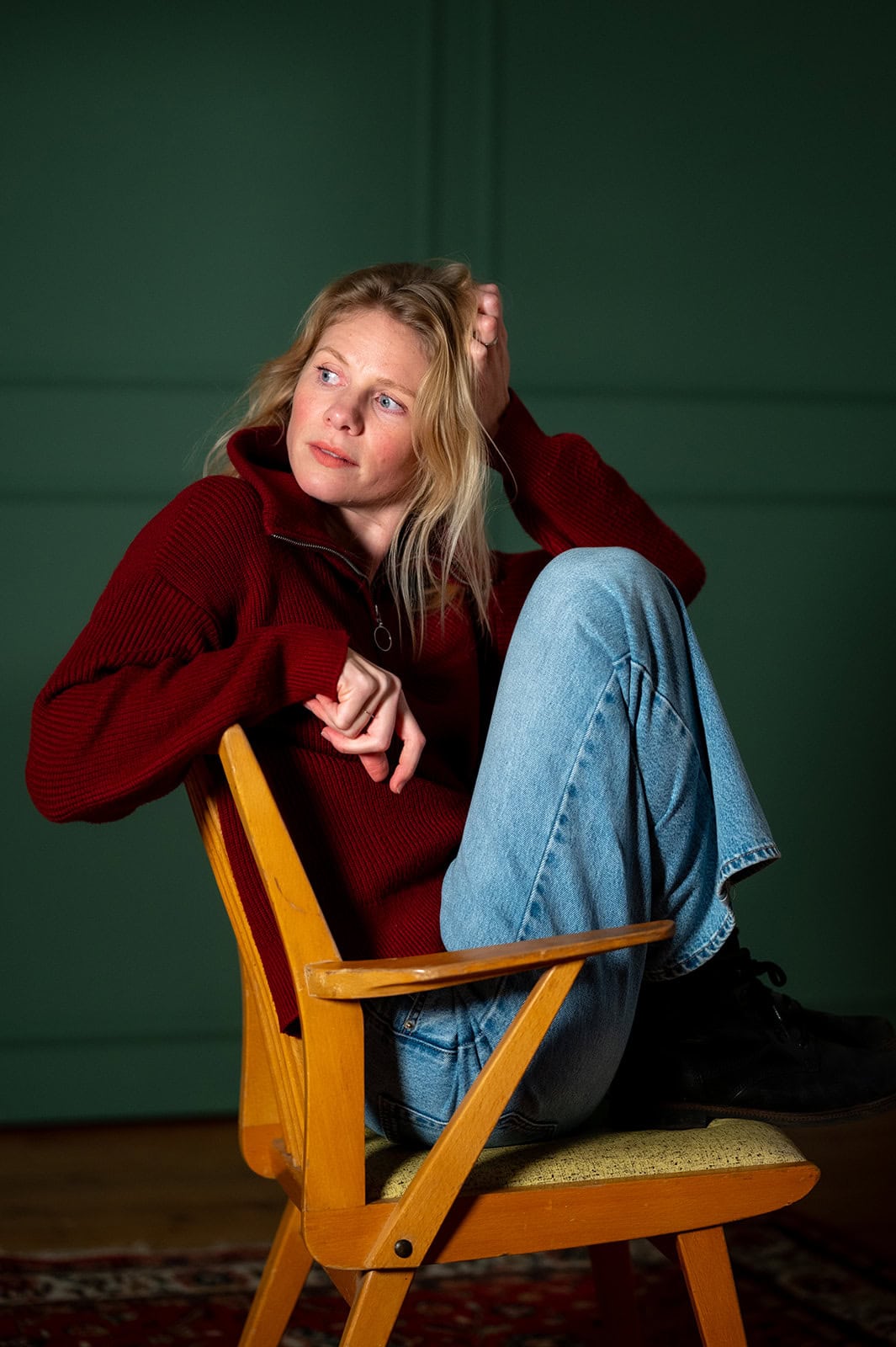 Junge Frau sitzt nachdenklich auf einem Holzstuhl vor grünem Hintergrund.