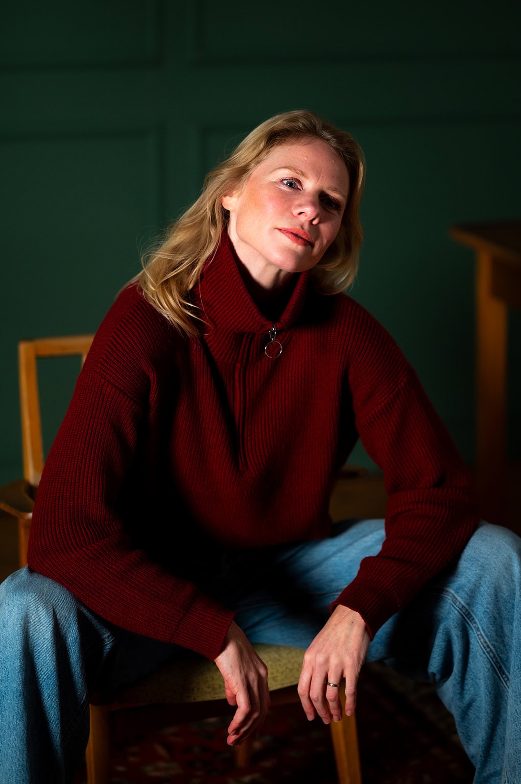 Nachdenkliche Frau in rotem Pullover sitzt auf einem Stuhl, grüner Hintergrund.