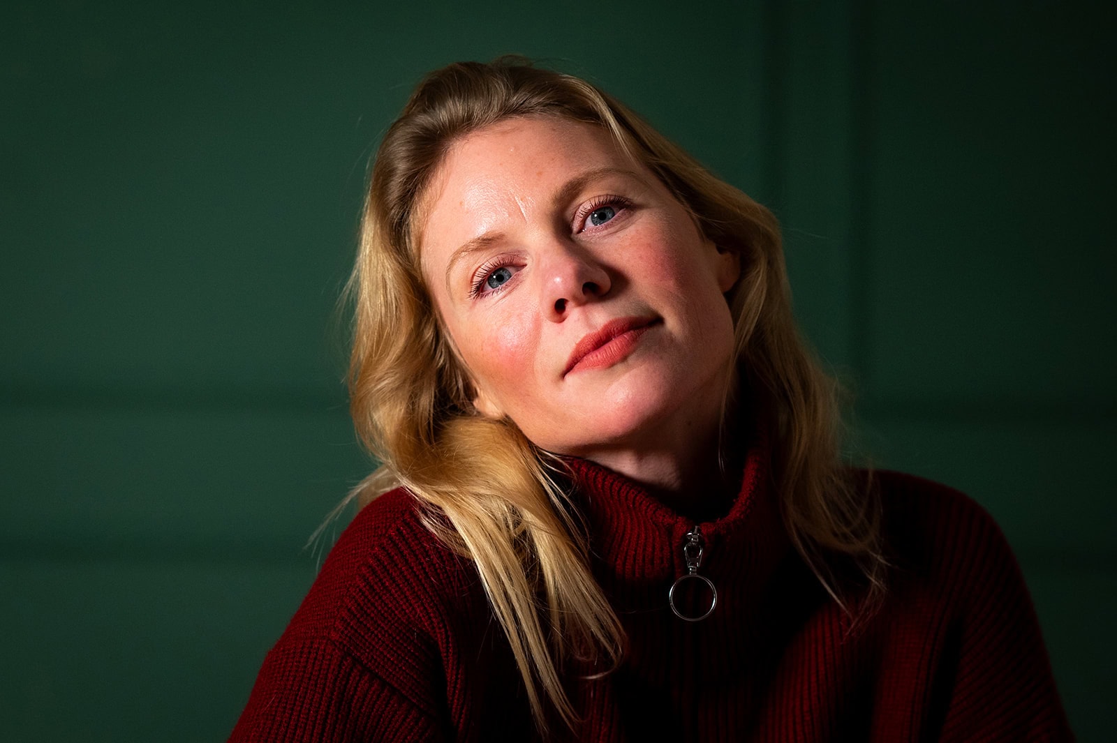 Junge Frau mit blondem Haar und nachdenklichem Blick, trägt einen dunklen Pullover vor grünem Hinter.